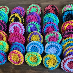 May include: A collection of 20 colorful crocheted scrubbers in various colors, including pink, blue, green, yellow, purple, and black. The scrubbers are arranged in rows on a wooden surface.