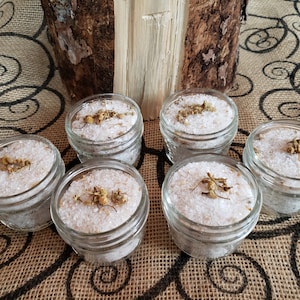 Op de afbeelding: Zes kleine, heldere glazen potjes gevuld met witte badzouten en gegarneerd met gedroogde bloemen. De potjes zijn gerangschikt op een jute oppervlak met een decoratief wervelpatroon. Een stuk hout staat op de achtergrond.