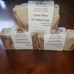 May include: Three bars of handmade soap with a brown and white swirl pattern. The soap is labeled "Soaps & More Natural Skincare Since 2006 Cocoa Honey & Oatmeal Soap" with a net weight of 4.5 oz.