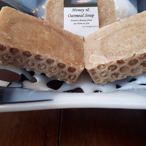 May include: Two bars of honey and oatmeal soap on a white and black plate. The soap is a light brown color and has a honeycomb design. The label reads "Honey & Oatmeal Soap, Nature's Bounty from our farm to you, Net Weight 4.5 oz."