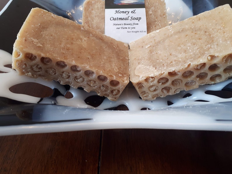 May include: Two bars of honey and oatmeal soap on a white and black plate. The soap is a light brown color and has a honeycomb design. The label reads "Honey & Oatmeal Soap, Nature's Bounty from our farm to you, Net Weight 4.5 oz."