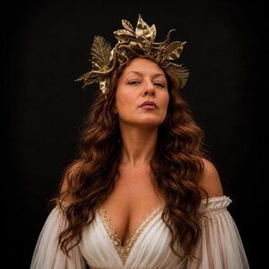 May include: A gold-colored headpiece featuring intricate metallic leaves and fern-like fronds. The accessory is worn atop long, wavy brown hair, complementing a white off-the-shoulder dress with gold lace trim along the neckline and waist.