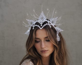 May include: A white headpiece featuring intricate lace floral motifs, delicate beaded accents, and tall, spiky crystalline structures. The design incorporates small hanging pearl-like beads along the scalloped edge, creating a crown-like appearance.