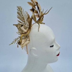 May include: A gold-colored headpiece with a snake and leaf design. The headpiece is made of metal and is worn on a mannequin's head.