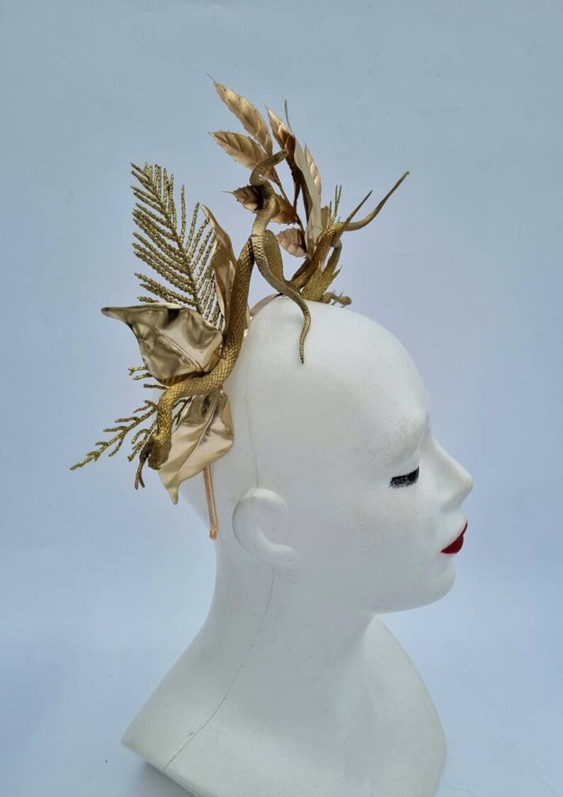 May include: A gold-colored headpiece with a snake and leaf design. The headpiece is made of metal and is worn on a mannequin's head.