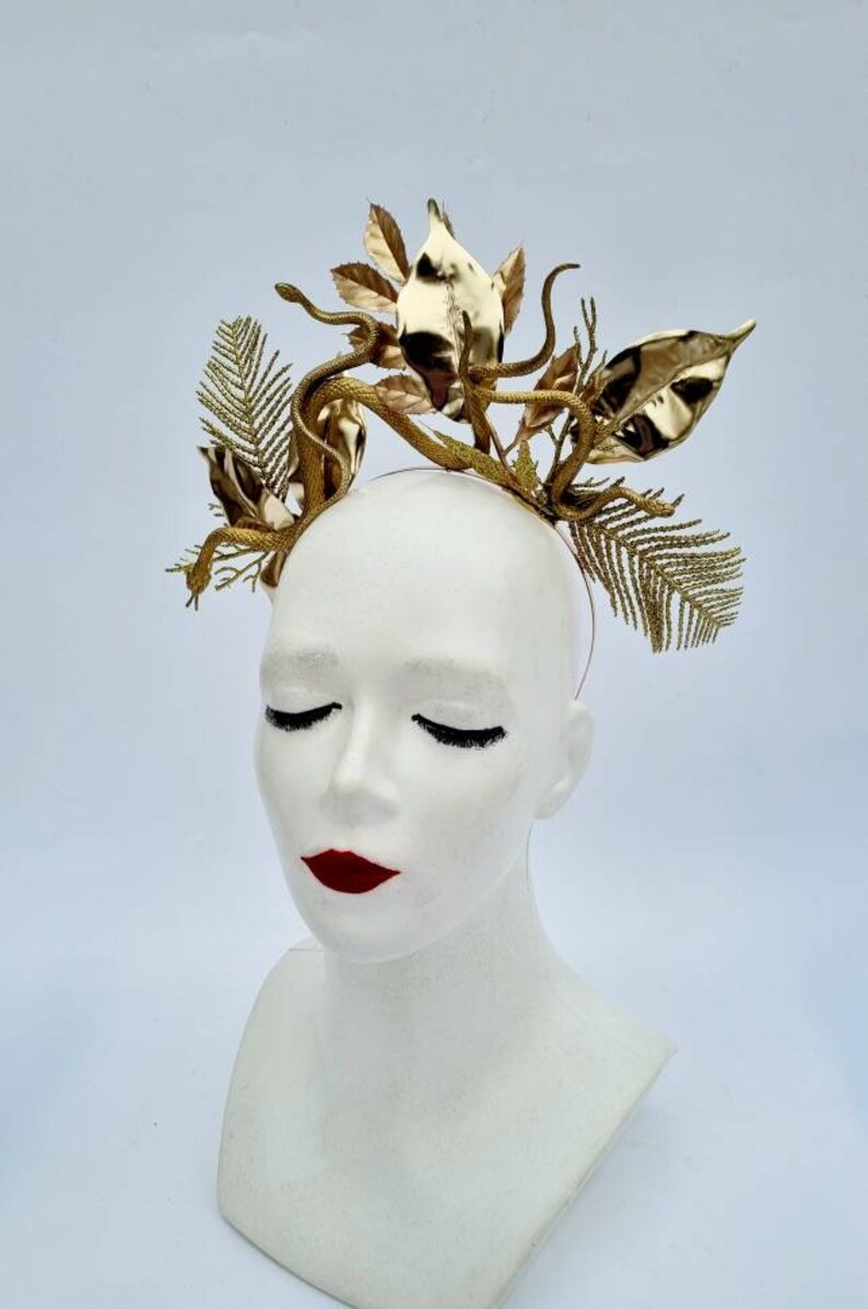May include: A gold headband with a snake and leaf design. The headband is made of metal and has a shiny finish.