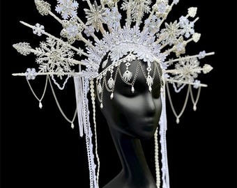 May include: A white, ornate headpiece with snowflake and floral designs. The headpiece features dangling chains and beaded accents. It is displayed on a black mannequin head against a black background. The headpiece is likely for a costume or special occasion.