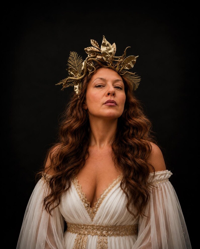 May include: A gold-colored headpiece featuring intricate metallic leaves and fern-like fronds. The accessory is worn atop long, wavy brown hair, complementing a white off-the-shoulder dress with gold lace trim along the neckline and waist.
