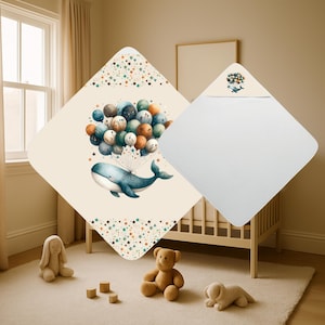 Op de afbeelding: Een babydeken met een grillig ontwerp met een blauwe walvis die zweeft met kleurrijke ballonnen. De deken heeft een crèmekleurige achtergrond met een sterrenpatroonrand. Een bijpassende badhanddoek met capuchon is ook zichtbaar.