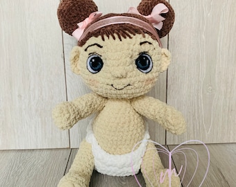 May include: A crocheted doll with brown hair, wearing a white nappy and a pink headband with bows. The doll has large blue eyes and a smiling face.