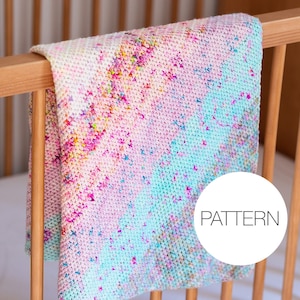 Op de afbeelding: Een handgemaakte babydeken met een kleurrijk, gespikkeld ontwerp. De deken is over een houten spijlen van een ledikant gedrapeerd. Het kleurenpalet omvat roze, blauw en geel. Het woord "PATTERN" staat in een witte cirkel.