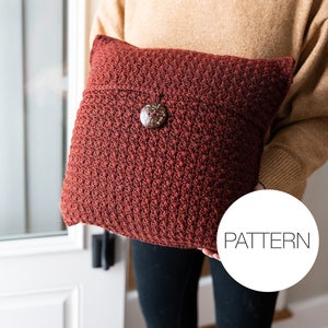 May include: A red crocheted pillow with a wooden button. The pillow is being held by a person wearing a tan sweater and black pants. The text "PATTERN" is visible in a white circle.