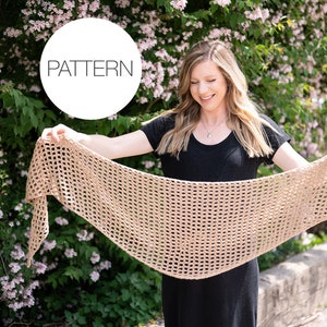 May include: A woman wearing a black dress holds a beige crochet shawl in front of her. The shawl has a lacy, openwork design. The word "PATTERN" is written in a white circle in the upper left corner of the image.