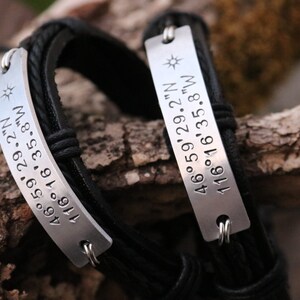 May include: Two black leather bracelets with silver rectangular tags. Each tag is engraved with coordinates and a sunburst symbol. The bracelets are displayed on a wooden surface, showcasing a rustic aesthetic.