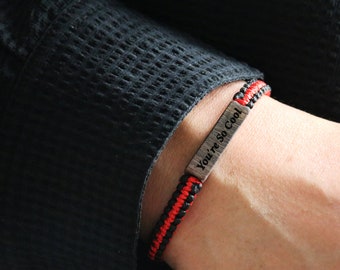 May include: A black and red braided bracelet with a silver metal tag that reads "You're So Cool".