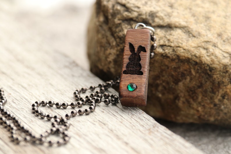 Peut inclure: Un collier avec un pendentif en bois sculpt&eacute; en forme de lapin et une pierre pr&eacute;cieuse verte. Le collier a une cha&icirc;ne en m&eacute;tal noir.
