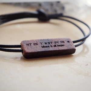 Peut inclure: Un bracelet en cordon noir avec un élément central rectangulaire en bois. Le bois est gravé de coordonnées et de la phrase "Where it all began". Le bracelet se ferme par un nœud.