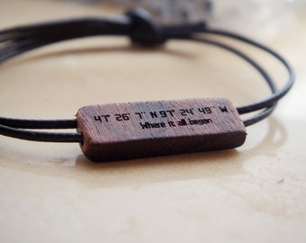 May include: A black cord bracelet with a rectangular wooden centerpiece. The wood is engraved with coordinates and the phrase "Where it all began." The bracelet is tied with a knot closure.