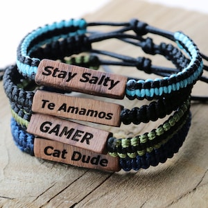 Może przedstawiać: Cztery plecione bransoletki z drewnianymi zawieszkami. Zawieszki mają następujący tekst: Stay Salty, Te Amamos, GAMER, Cat Dude.