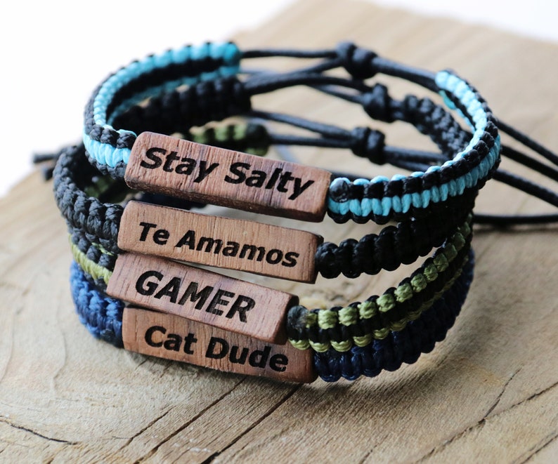 Może przedstawiać: Cztery plecione bransoletki z drewnianymi zawieszkami. Zawieszki mają następujący tekst: Stay Salty, Te Amamos, GAMER, Cat Dude.