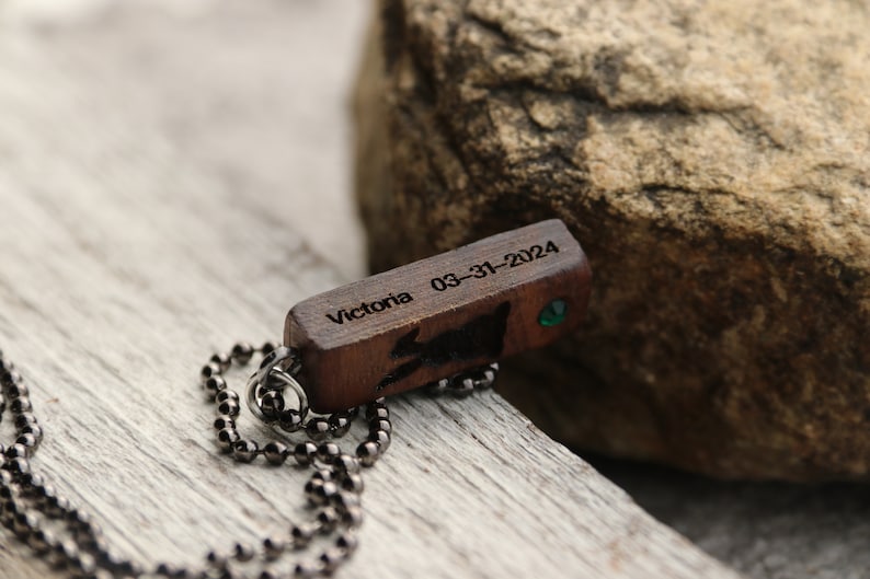 Peut inclure: Un collier avec un pendentif en bois et une cha&icirc;ne noire. Le pendentif est grav&eacute; avec le texte "Victoria 03-31-2024" et comporte une petite pierre pr&eacute;cieuse verte.