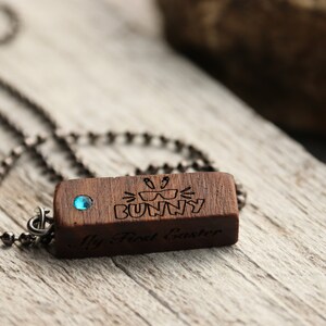 Peut inclure: Un collier avec un pendentif en bois et une pierre pr&eacute;cieuse bleue. Le pendentif est grav&eacute; des mots "My First Easter" et un lapin portant des lunettes de soleil.