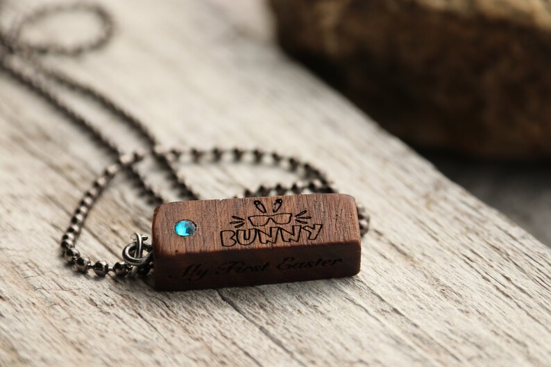 Peut inclure: Un collier avec un pendentif en bois et une pierre pr&eacute;cieuse bleue. Le pendentif est grav&eacute; des mots "My First Easter" et un lapin portant des lunettes de soleil.
