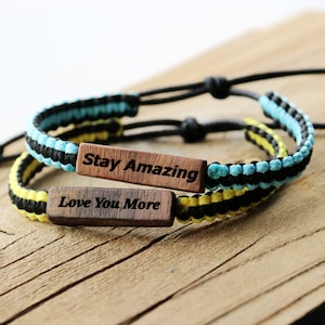 Może przedstawiać: Dwie bransoletki przyjaźni z czarnymi, ż&oacute;łtymi i niebieskimi plecionymi sznurkami. Jedna bransoletka ma drewniany znaczek z napisem "Stay Amazing", a druga drewniany znaczek z napisem "Love You More".