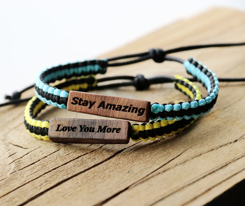 Może przedstawiać: Dwie bransoletki przyjaźni z czarnymi, ż&oacute;łtymi i niebieskimi plecionymi sznurkami. Jedna bransoletka ma drewniany znaczek z napisem "Stay Amazing", a druga drewniany znaczek z napisem "Love You More".