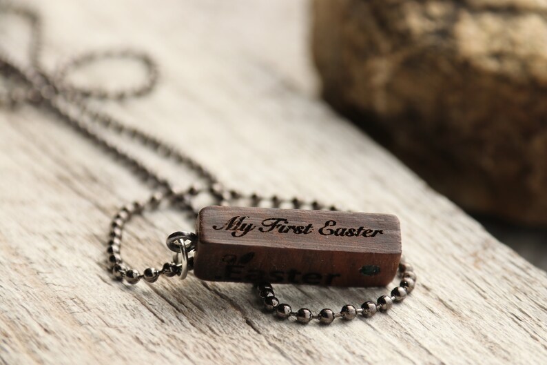 Peut inclure: Un collier avec un pendentif en bois et une cha&icirc;ne noire. Le pendentif est grav&eacute; avec les mots "My First Easter".