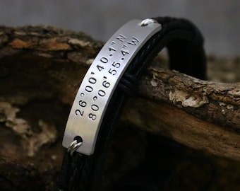 May include: A black leather bracelet with a silver plate engraved with the coordinates "26°00'40.1"N 80°06'55.4"W".