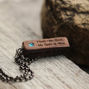 Peut inclure: Un collier avec un pendentif en bois et une pierre pr&eacute;cieuse bleue. Le pendentif est grav&eacute; avec le texte "Flesh Has Died, My Spirit Is Alive" et la date "03-11-2021". Le collier est sur une cha&icirc;ne noire.