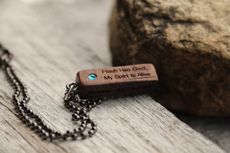 Peut inclure: Un collier avec un pendentif en bois et une pierre pr&eacute;cieuse bleue. Le pendentif est grav&eacute; avec le texte "Flesh Has Died, My Spirit Is Alive" et la date "03-11-2021". Le collier est sur une cha&icirc;ne noire.