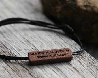 May include: A black cord bracelet with a rectangular wooden charm. The charm is engraved with coordinates and the phrase "Where it all began." The bracelet is displayed on a weathered wooden surface.