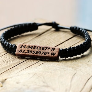 May include: A black braided bracelet with a wooden tag engraved with the coordinates "34.9451167&deg; N -82.3953976&deg; W".