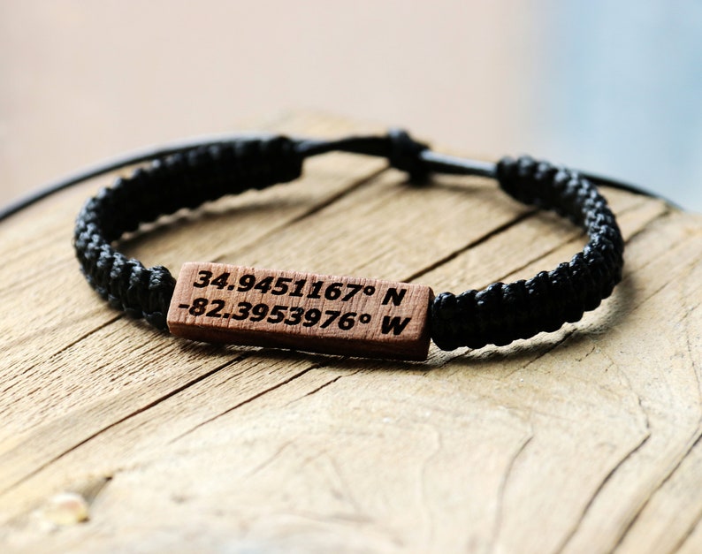 May include: A black braided bracelet with a wooden tag engraved with the coordinates "34.9451167&deg; N -82.3953976&deg; W".