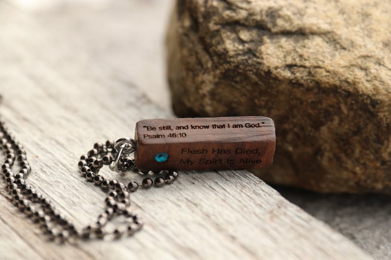 Peut inclure: Un collier avec un pendentif en bois et une pierre pr&eacute;cieuse bleue. Le pendentif porte le texte "Sois tranquille, et sache que je suis Dieu." Psaume 46:10 et "La chair est morte, mon esprit est vivant".