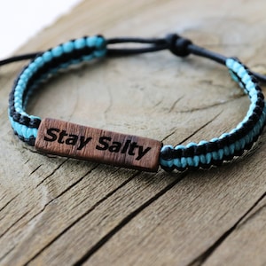 Może przedstawiać: Bransoletka pleciona na niebiesko i czarno z drewnianym wisiorkiem z napisem "Stay Salty".