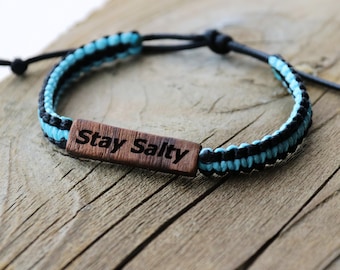 May include: A blue and black braided bracelet with a wooden charm that says "Stay Salty".
