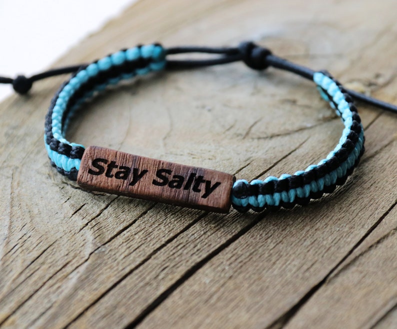 Może przedstawiać: Bransoletka pleciona na niebiesko i czarno z drewnianym wisiorkiem z napisem "Stay Salty".