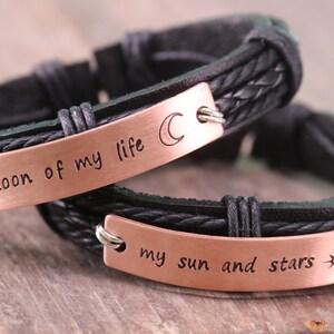 May include: Two black leather bracelets with copper-colored metal plates. The top bracelet has the text "moon of my life" and a crescent moon symbol. The bottom bracelet has the text "my sun and stars" and a sun with stars symbol.