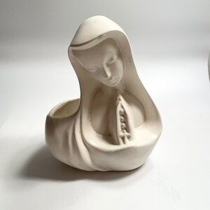 Puede incluir: Macetero de cerámica blanca con forma de mujer en actitud de oración. El macetero tiene un acabado liso y mate y una superficie ligeramente texturizada.