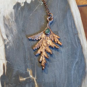 May include: A copper-colored leaf pendant necklace with a small, oval, green gemstone set near the top. The leaf has detailed veining and is suspended from a delicate chain. The necklace is displayed on a gray, textured surface.