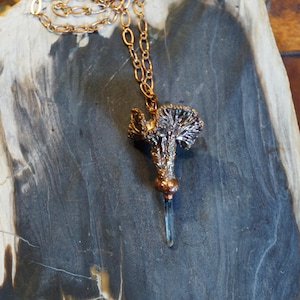 Puede incluir: Un collar de tono cobre con un colgante texturizado y un cristal azul. El colgante tiene un diseño similar a un árbol. El collar se muestra sobre una superficie gris y marrón.