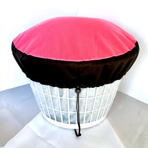 May include: A white laundry basket with a pink and black cover. The cover is round and fits over the top of the basket. The cover has a drawstring closure. The basket is made of a white plastic material.
