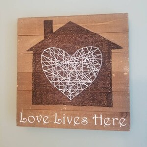 May include: Wooden wall art featuring a house silhouette with a string art heart. The house is dark brown, and the heart is made of white string. The words "Love Lives Here" are written in white script at the bottom.