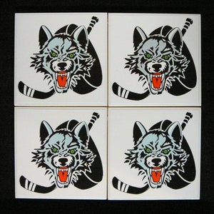 May include: Four white ceramic coasters with a black and white image of a wolf's head with a hockey stick. The wolf's eyes are green and its mouth is open, showing sharp teeth.