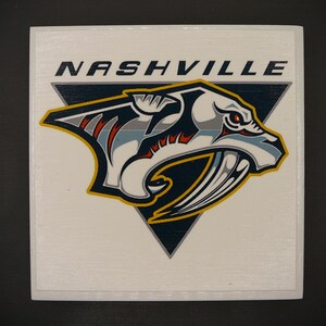 May include: A white square sticker with the Nashville Predators NHL team logo. The logo features a stylized saber-tooth tiger with a blue, white, and red striped mane, a yellow and black saber tooth, and a red eye. The text "NASHVILLE" is printed in black at the top of the sticker.