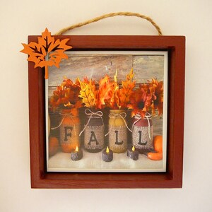 May include: A framed picture with a rustic wood background featuring four mason jars decorated with burlap and the word "FALL" in black letters. The jars are filled with fall leaves and surrounded by small candles.