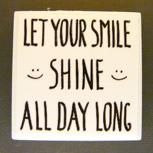 May include: White wooden sign with black text that reads "Let your smile shine all day long". Two smiley faces are drawn on either side of the word "shine".